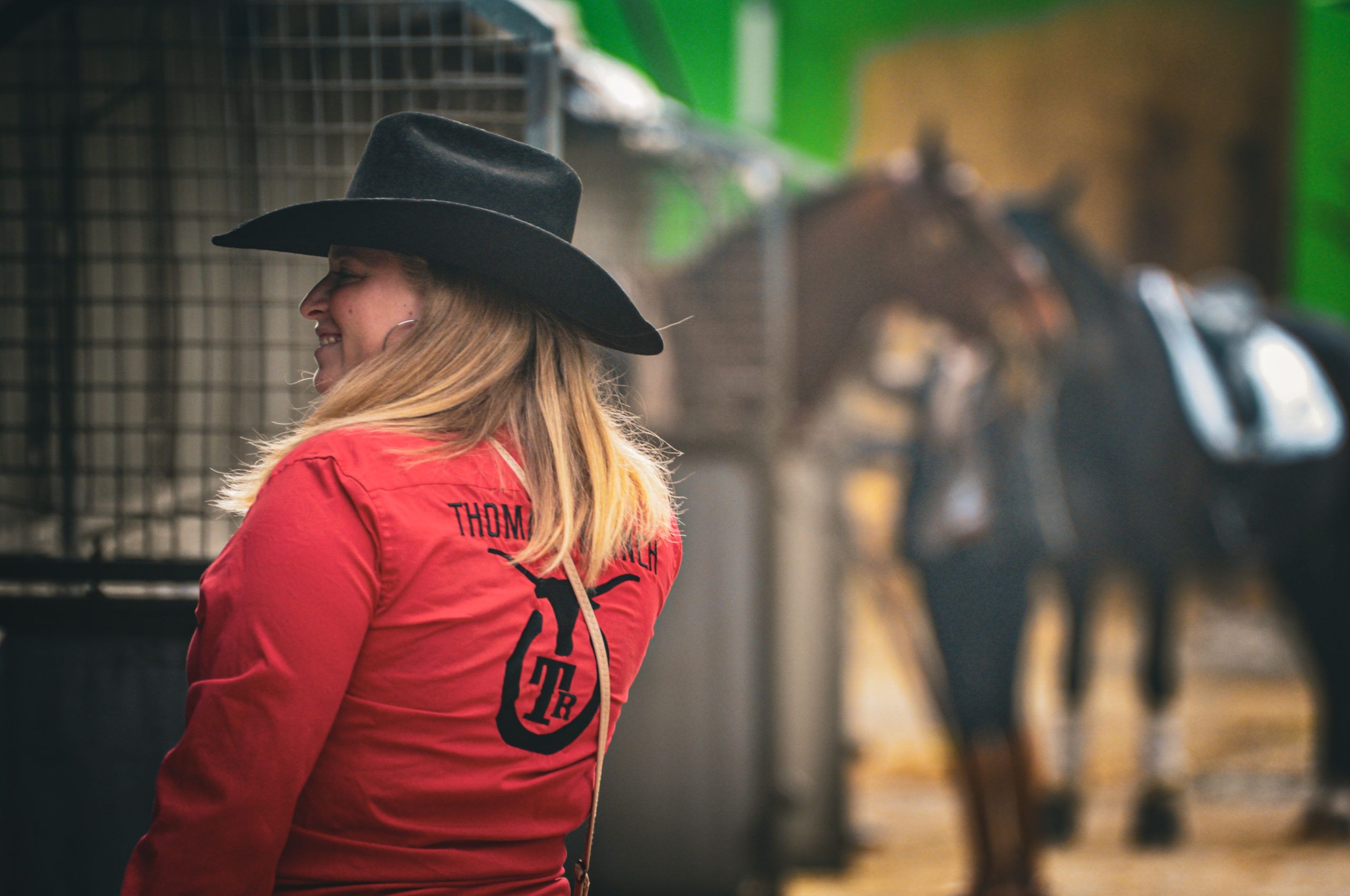 Cavalière de dos en chemise western rouge brodée « Thomas' Ranch » et du logo TR, chapeau noir, chevaux en fond