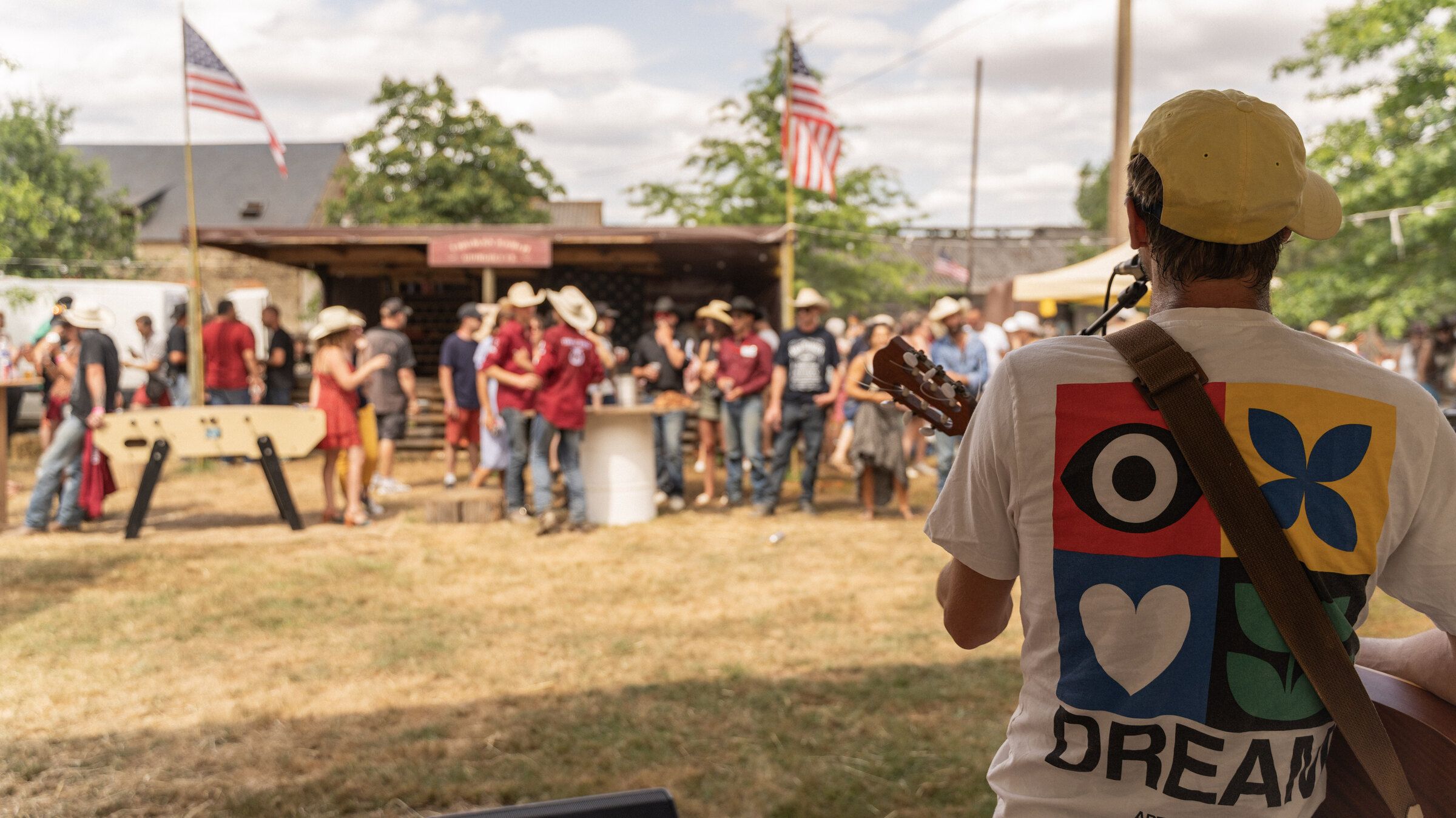 Chanteur à la guitare vu de dos, face au public du festival rassemblé devant le bar, drapeaux américains