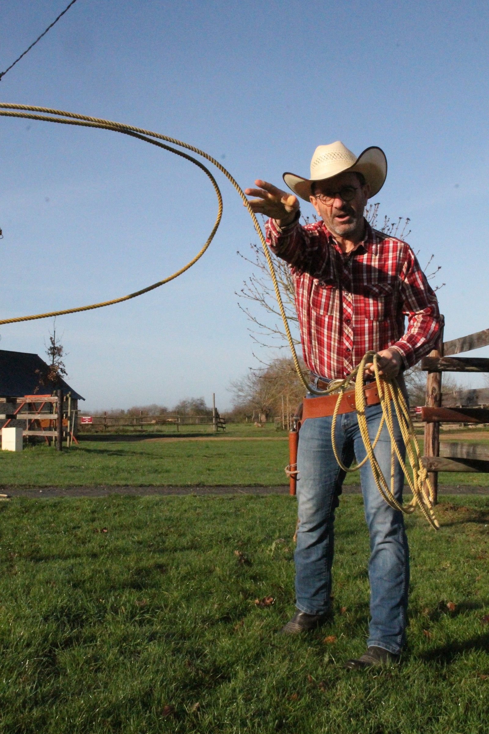 Jacky Thomas fait tourner un lasso dans un pré du ranch, en chemise à carreaux et chapeau de cow-boy