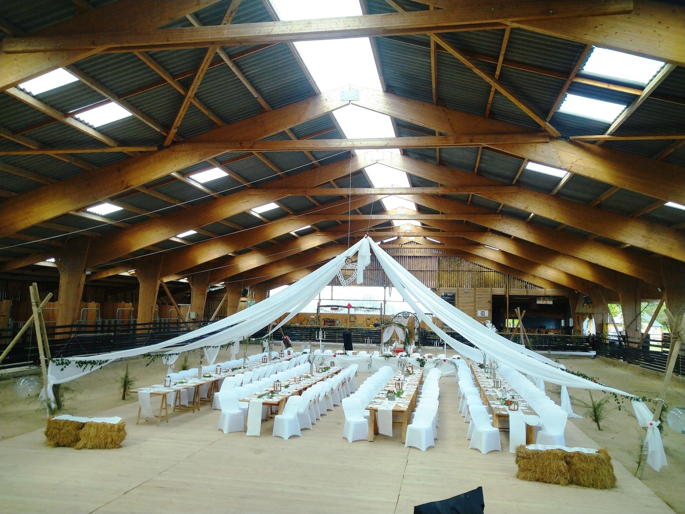Le manège du ranch dressé pour un mariage : voilages blancs tendus sous la charpente, longues tables et bottes de paille
