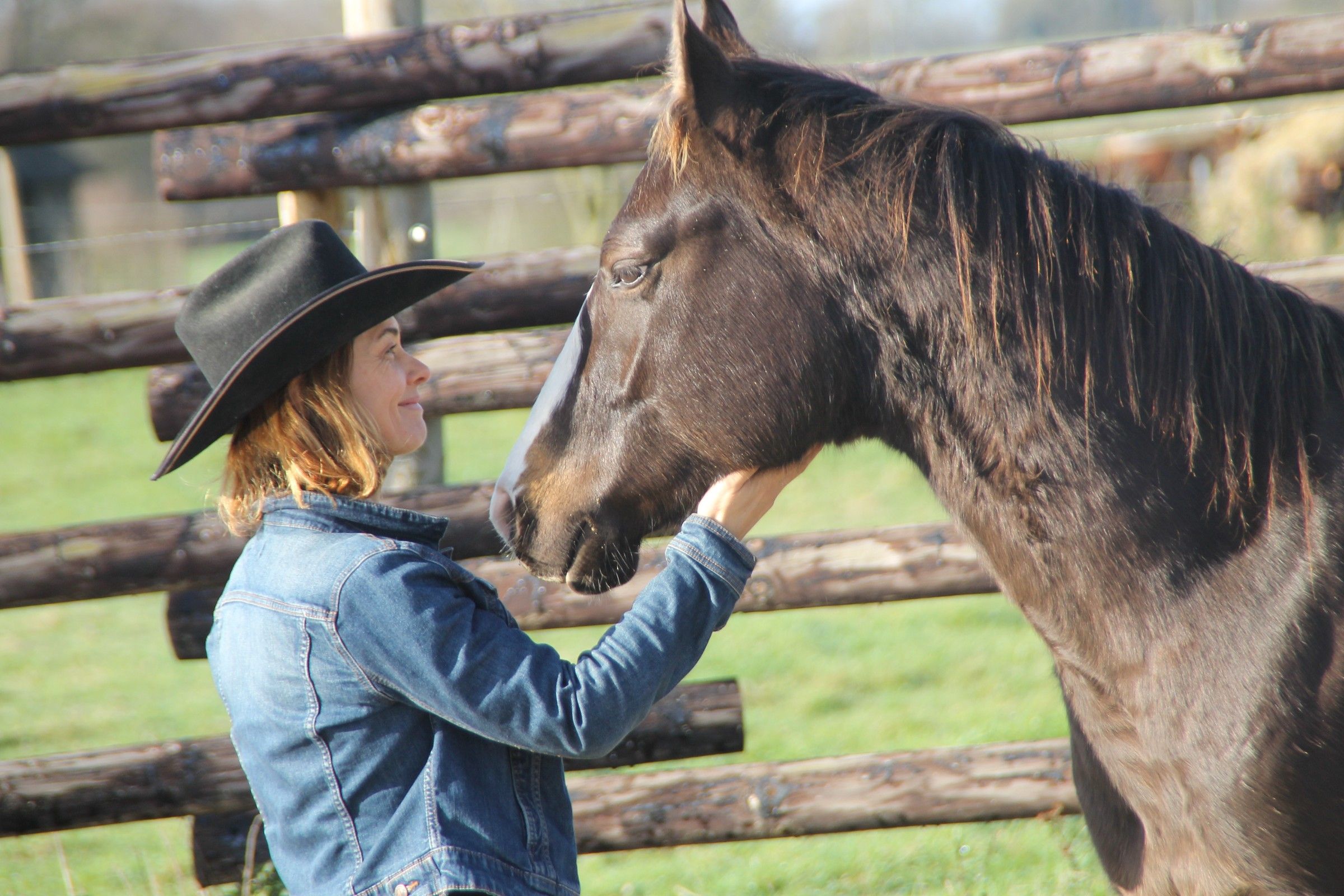 Stéphanie Thomas, en chapeau de cow-boy, caresse un cheval bai à la barrière en rondins du ranch